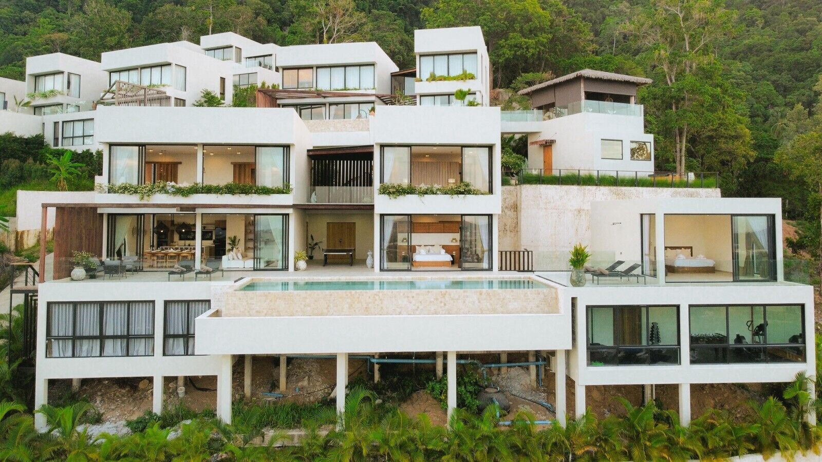 A modern multi-story residential building with a central swimming pool, balconies with plants, and a lush green backdrop.
