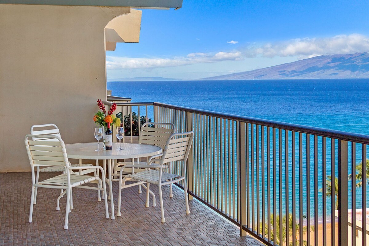 Ein Balkon mit Tisch und Stühlen, mit Blick auf ein Gewässer und eine gebirgige Landschaft.