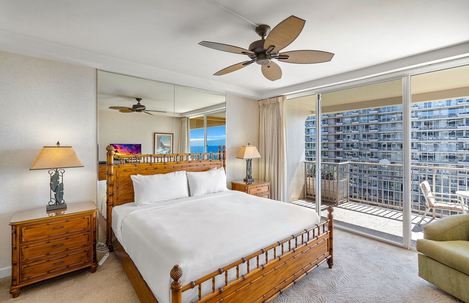 Quarto de hotel com uma cama grande, duas mesas de cabeceira com luminárias, um ventilador de teto, uma janela ampla com vista para um edifício e uma varanda com cadeiras.
