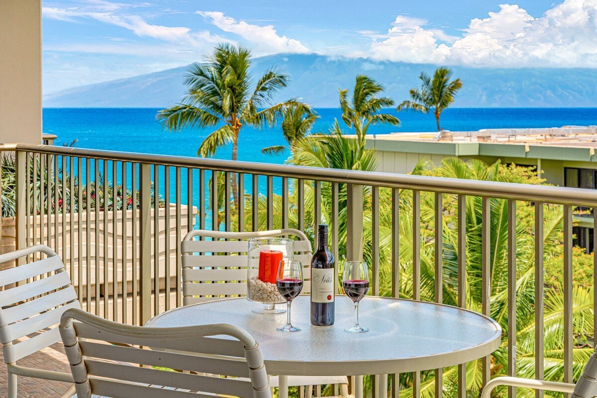Ein Balkon mit Tisch für zwei Personen, ausgestattet mit einer Weinflasche und Gläsern, mit Blick auf einen Strand mit Palmen und einem Berg in der Ferne.