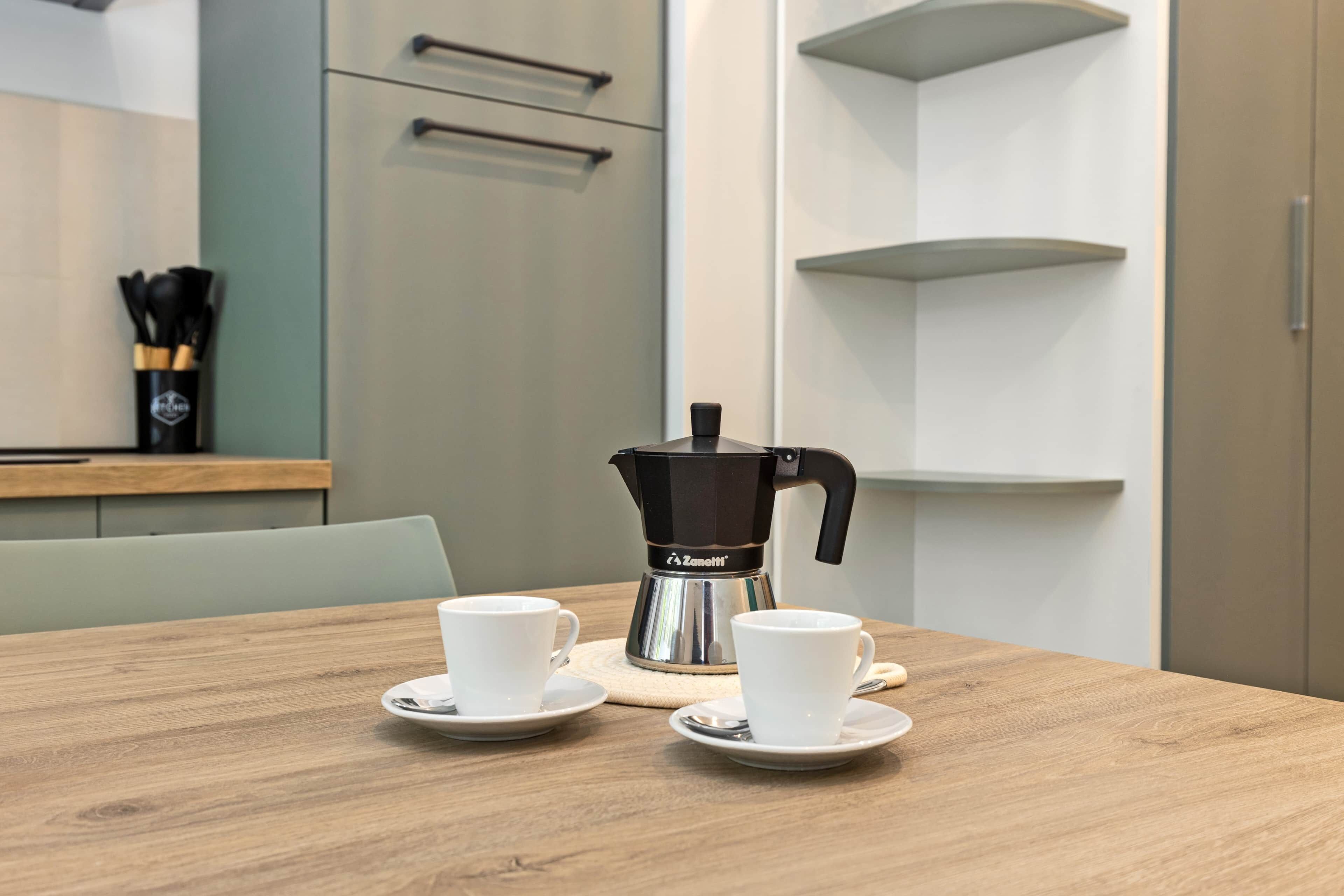 A modern kitchen with a coffee maker on the counter, two white cups on saucers, and a wooden table.