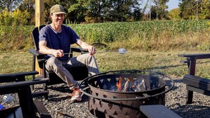 A person sitting by a fire pit with a metal grill, surrounded by outdoor furniture and a natural landscape.