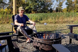 A person sitting by a fire pit with a metal grill, surrounded by outdoor furniture and a natural landscape.