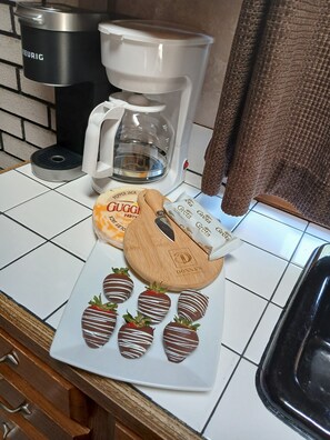 A kitchen counter with a coffee maker, a mug, a plate with chocolate-covered strawberries, and a wooden cookie cutter. - Romantic Queen Cottage with Jacuzzi & Fireplace (Berlin)