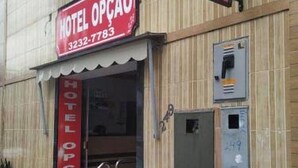 A hotel entrance with a red awning displaying the name 'HOTEL OPOCAO' and a phone number. There are steps leading up to the entrance, and a utility box is mounted on the wall to the right. - Hotel Opção (Manaus)