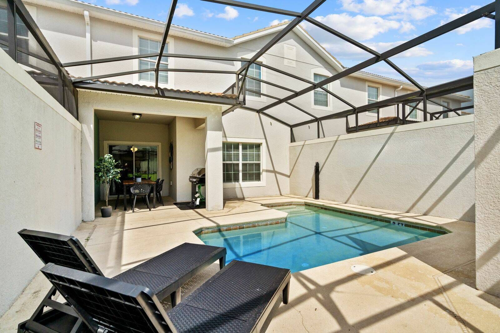 Une piscine entourée de parois en verre, un espace patio avec une table et des chaises, et une vue sur un immeuble d’habitation en arrière-plan.