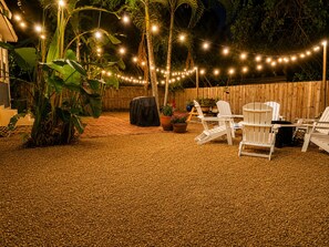 Un espace extérieur aménagé en terrasse, avec des guirlandes lumineuses, des fauteuils Adirondack blancs et des plantes en pot.