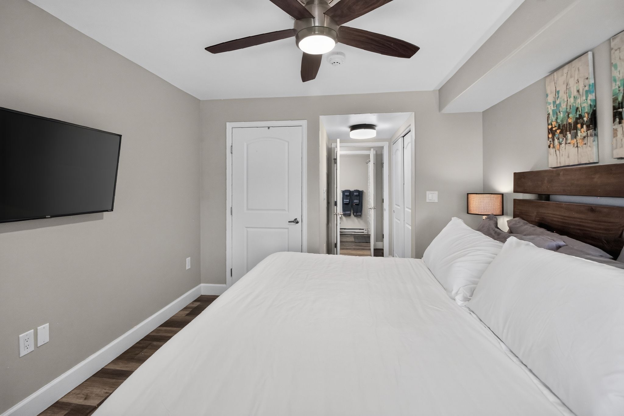 Une chambre à coucher comprenant un lit, un ventilateur de plafond, un téléviseur fixé au mur et une porte donnant accès à une salle de bain.