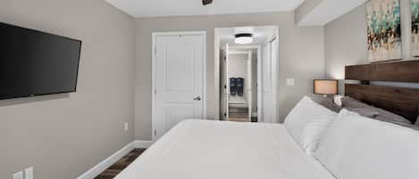 Une chambre à coucher comprenant un lit, un ventilateur de plafond, un téléviseur fixé au mur et une porte donnant accès à une salle de bain.