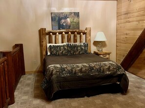 A bedroom with a wooden headboard, a bed with a camouflage-patterned bedspread, a nightstand with a lamp, and a painting of a moose on the wall. - The Rivercrest-Riverfront/Private Hot Tub/Deck (South Fork)