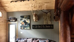 Een gezellige woonkamer met een leren bank, een gewei van een hert aan de muur, en een bordje met de tekst 'Life's Better In The Mountains'.