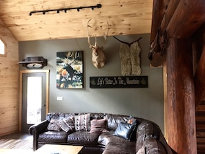 A cozy living room with a leather couch, a deer head mounted on the wall, and a sign that reads 'Life's Better In The Mountains.'