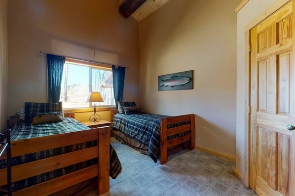 A room with two wooden beds, a window with curtains, a lamp, and a fish painting on the wall.