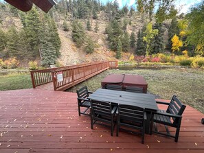 A wooden deck with a table and chairs, overlooking a forested area with a bridge and a pond.