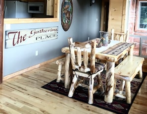 A rustic dining area with a wooden table and chairs, a sign reading 'The Gathering Place', and a kitchen in the background.