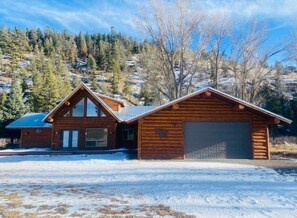 A log cabin with a garage, surrounded by snow-covered ground and a forested hillside.
