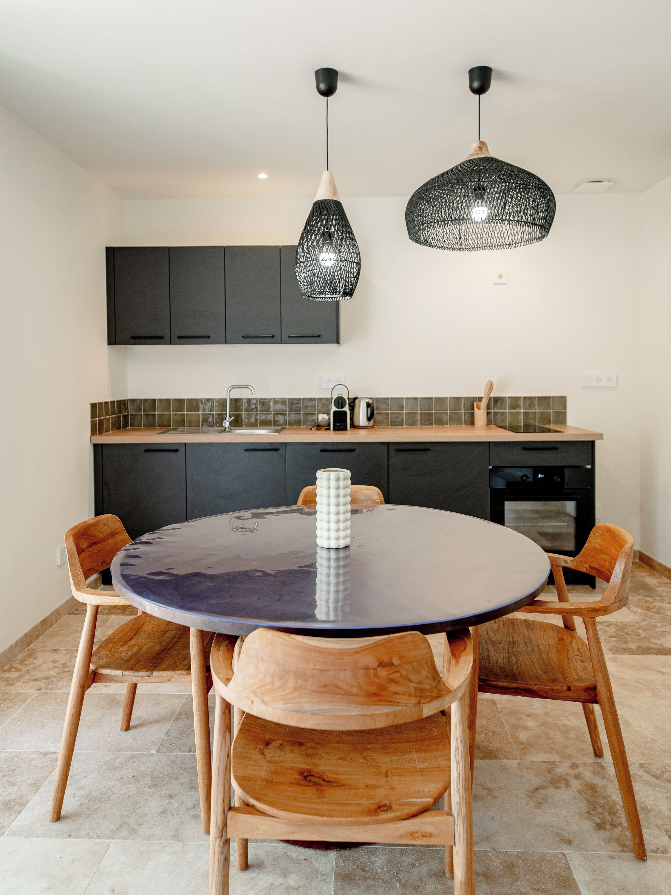 A modern kitchen with dark cabinets, a round table, wooden chairs, and two pendant lights.