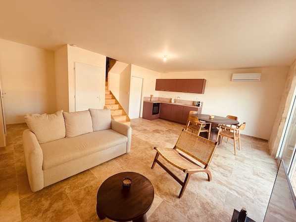 A living room with a sofa, a dining table with chairs, a kitchen area, and a staircase.