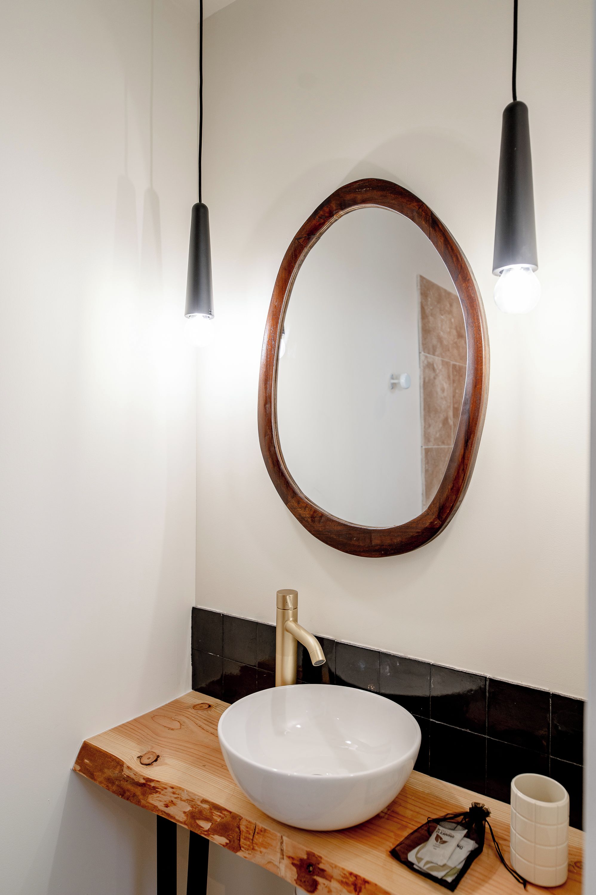 A bathroom with a wooden vanity, a white sink, a round mirror with a wooden frame, and two pendant lights.