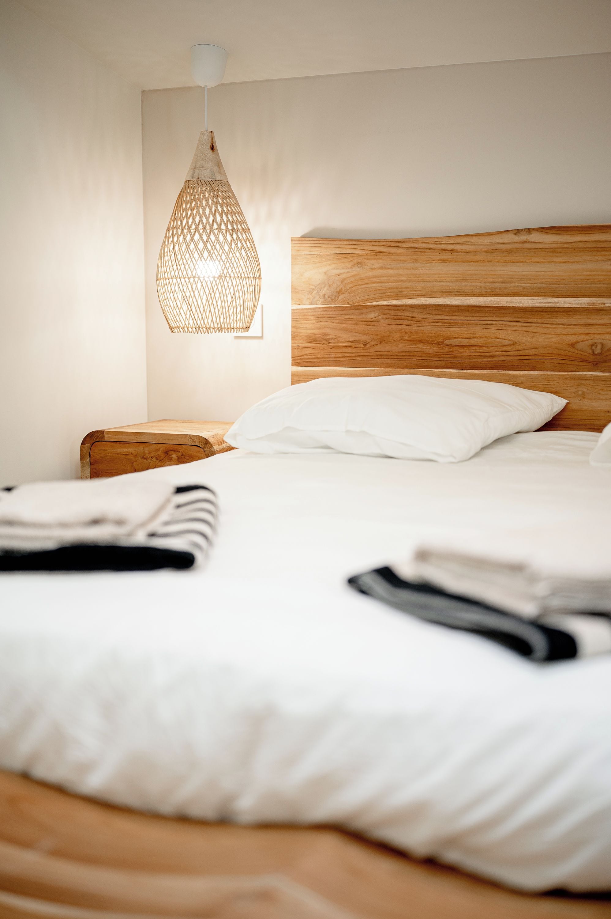 A neatly made bed with white linens, a wooden headboard, and a woven light fixture above it.