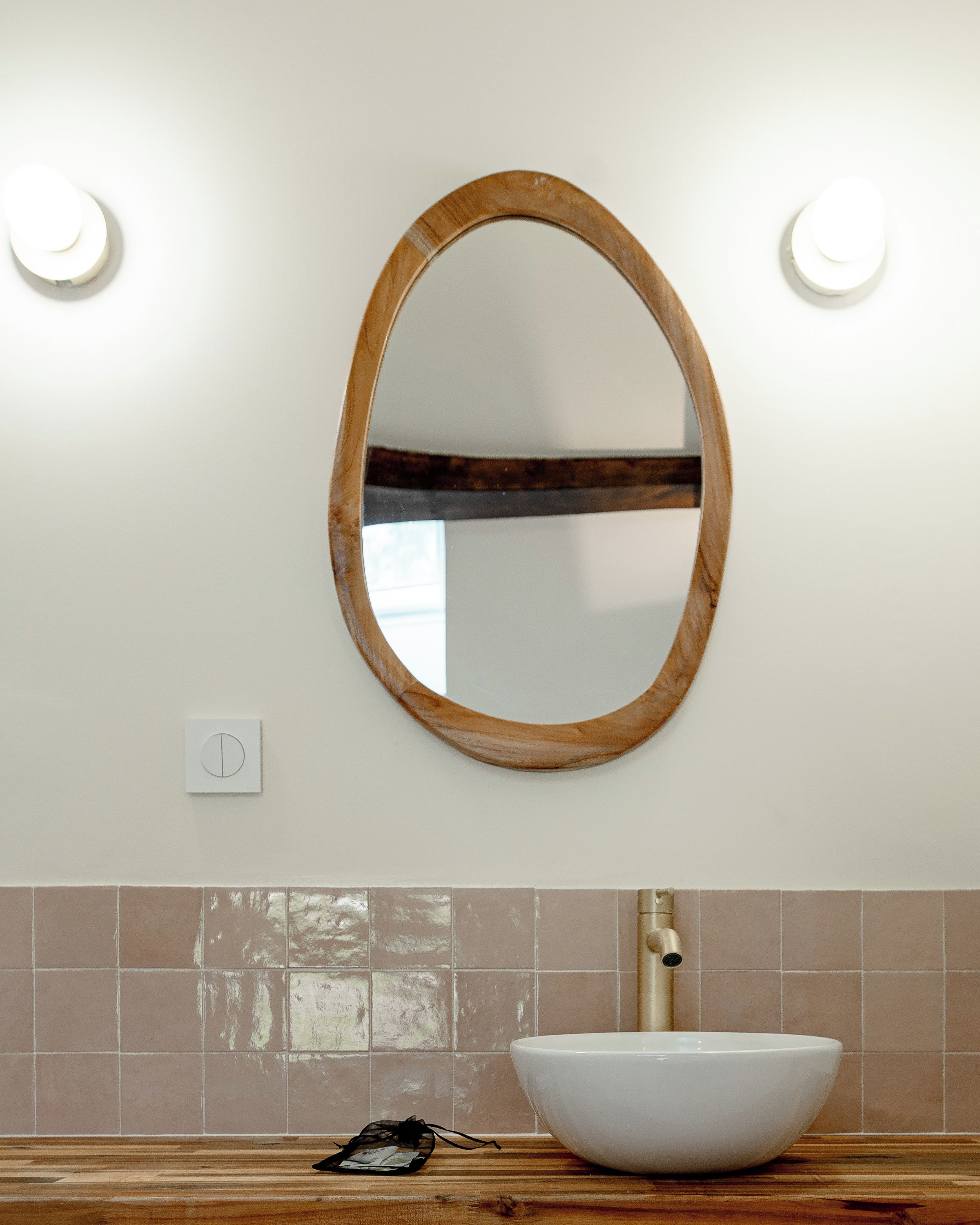 Une salle de bain avec un miroir ovale en bois, un lavabo blanc et un mur recouvert de carreaux.