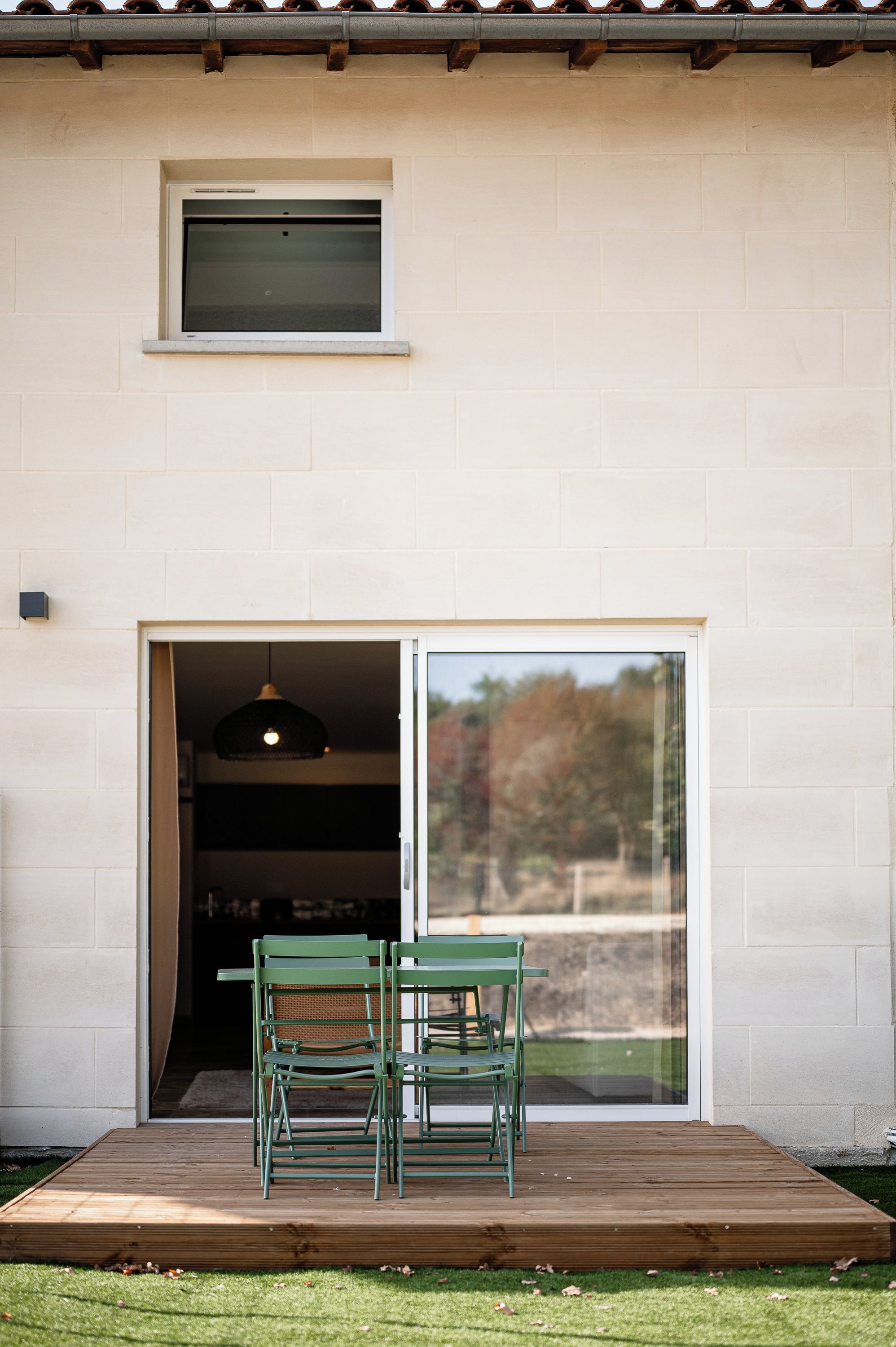 Entrada de edificio con puerta corredera de vidrio, una pequeña ventana arriba y una terraza de madera con dos sillas.