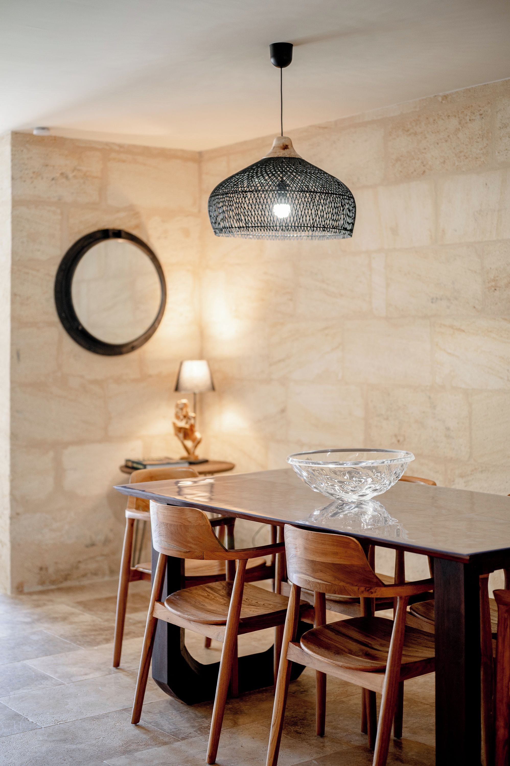 A dining area with a wooden table, chairs, a glass bowl, a hanging pendant light, a round mirror, and a small lamp on a side table.