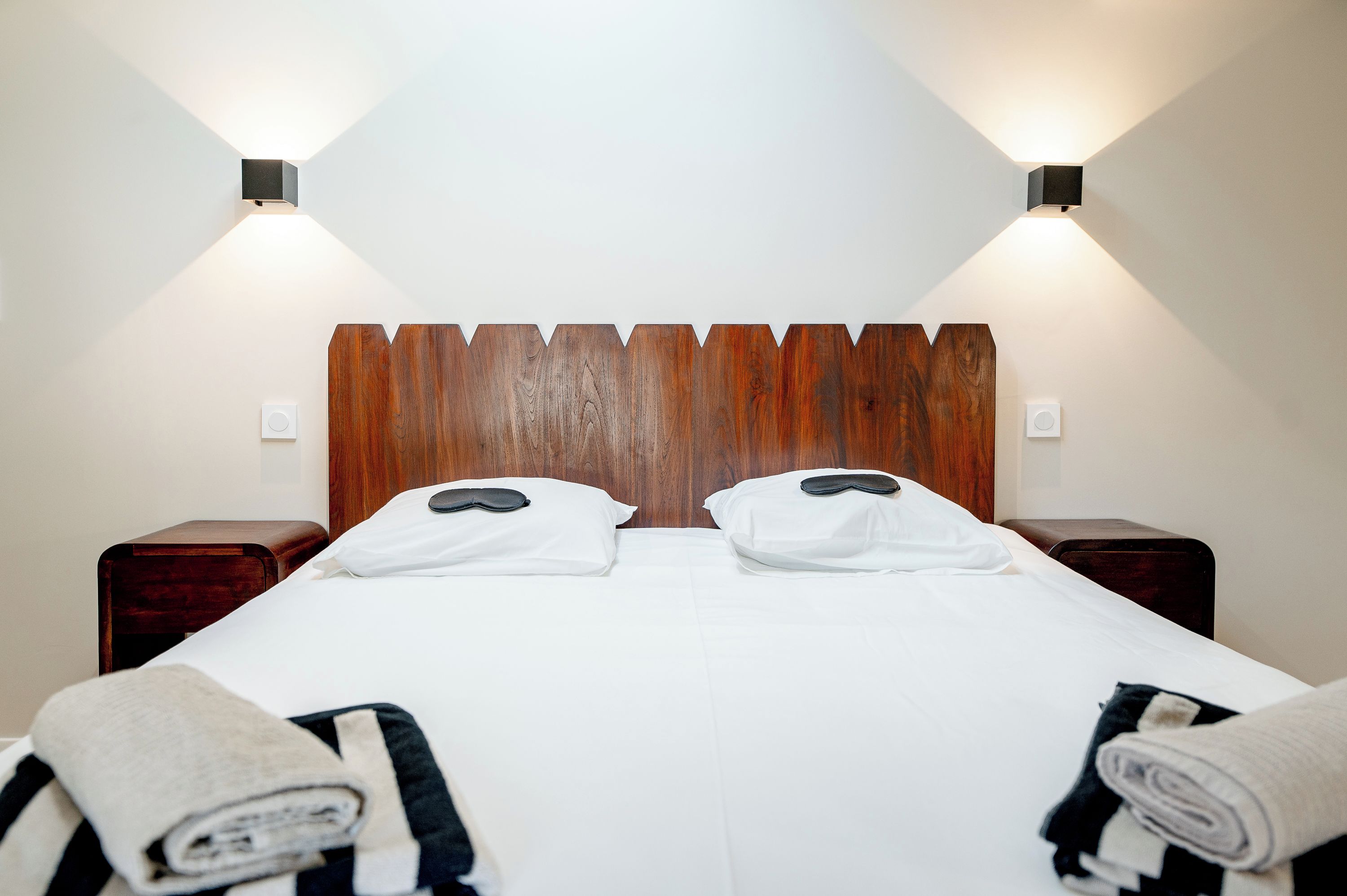 A neatly made bed with white linens, two pillows, and a wooden headboard. There are two bedside tables with lamps on either side.
