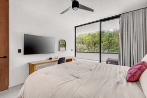 A modern bedroom with a large bed, a flat-screen TV mounted on the wall, a desk with a chair, and a view of trees through a sliding glass door.