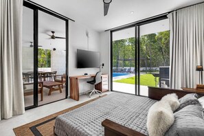 A bedroom with a large bed, a desk, and a TV. There is a sliding glass door leading to a patio with a pool and outdoor furniture.