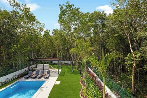Una piscina amb gandules, una zona de seients coberta i un jardí exuberant ple de plantes diverses.