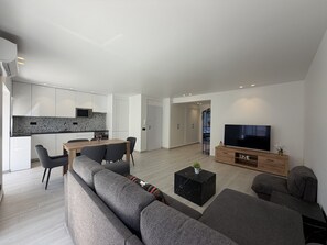 A modern living room with a sectional sofa, a dining area with a table and chairs, a kitchen with white cabinets, and a TV on a wooden stand. - Agua Plaza T2 With Pool AC (Albufeira)