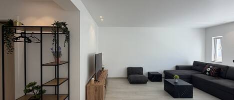 Ein modernes Wohnzimmer mit einem Ecksofa, einem Fernseher, einem Holztische und einem schwarzen Regal mit Pflanzen.