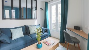 Una sala de estar moderna con un sofá azul, una mesa de centro con revistas, una planta en maceta, un escritorio con un radio y vistas al exterior a través de una ventana con cortinas.
