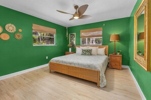 A room with a bed, a nightstand, a lamp, a ceiling fan, and a window with a view of greenery. - Tropicana Bungalow with Heated Pool, Pool Table & Tropical Vibes (Fort Myers)