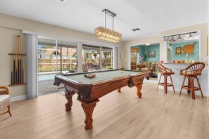 A pool table in a room with a view of a backyard, wooden flooring, and wicker furniture. - Tropicana Bungalow with Heated Pool, Pool Table & Tropical Vibes (Fort Myers)