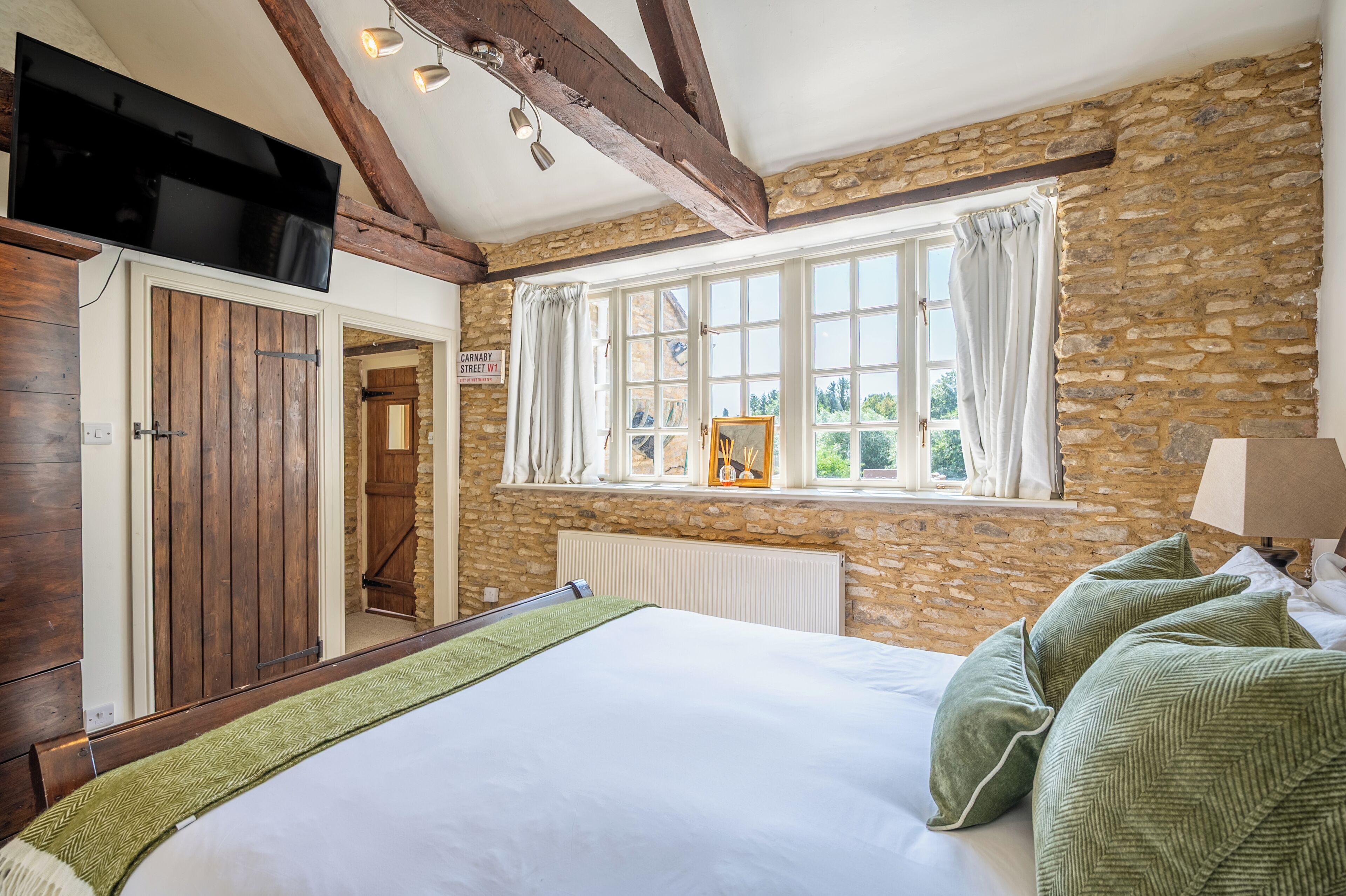 A bedroom with a stone wall, a bed with green pillows, a window with curtains, and a television mounted above the bed.