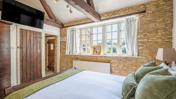 A bedroom with a stone wall, a bed with green pillows, a window with curtains, and a television mounted above the bed.