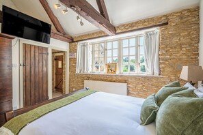 A bedroom with a stone wall, a bed with green pillows, a window with curtains, and a television mounted above the bed. - Gorgeous character Cottage on the edge of the Cotswolds (Middle Barton)