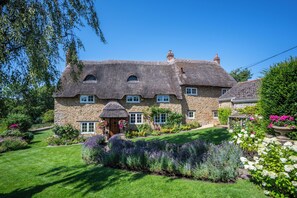 A stone house with a thatched roof, a small garden with lavender, and a clear blue sky.