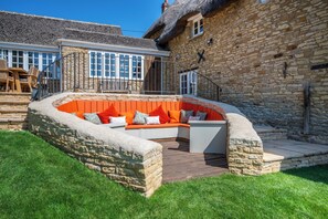 A stone building with a thatched roof, a stone bench with orange cushions, and a wooden deck. - Gorgeous character Cottage on the edge of the Cotswolds (Middle Barton)