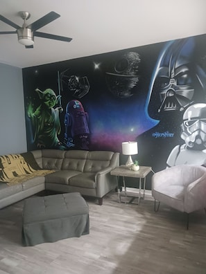 A living room with a Star Wars themed mural, a ceiling fan, a sofa, a small table with a lamp, and a chair. - Cozy 2-bedroom condo in peaceful Kissimmee with AC and gym (Kissimmee)