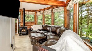 A cozy cabin interior with a leather sofa, a wood-burning stove, and large windows offering a forest view. - A Hexagonal Cabin in the Trees, Two Miles From Alyeska (Girdwood)