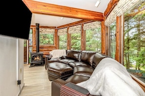 Ein gemütliches Blockhaus-Interieur mit Ledersofa, Kaminofen und großen Fenstern mit Blick auf den Wald.