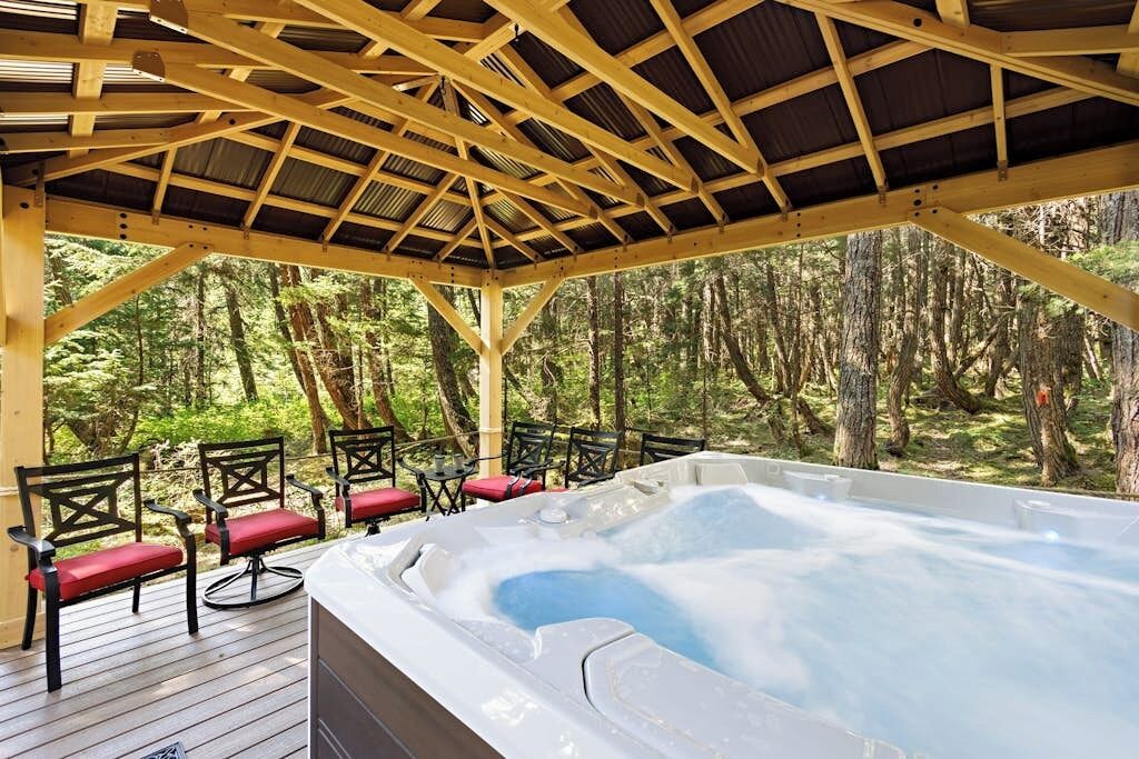 A hot tub under a wooden pergola with chairs, surrounded by a forest.