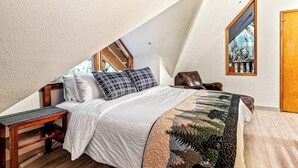 A bedroom with a sloped ceiling, a bed with a quilt, a bench, and a painting on the wall. - A Hexagonal Cabin in the Trees, Two Miles From Alyeska (Girdwood)