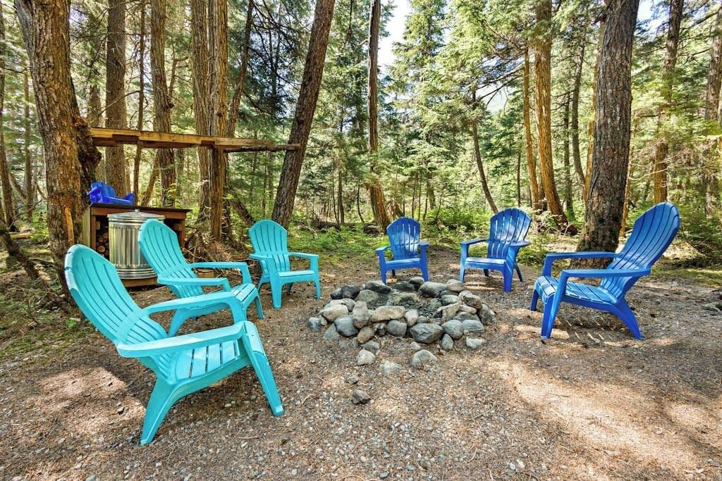 A forested campsite with several Adirondack chairs arranged around a fire pit.