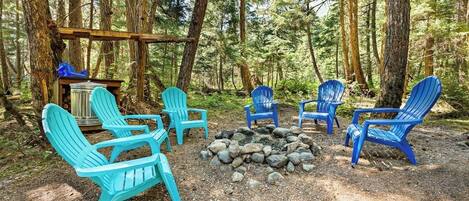 A forested campsite with several Adirondack chairs arranged around a fire pit.
