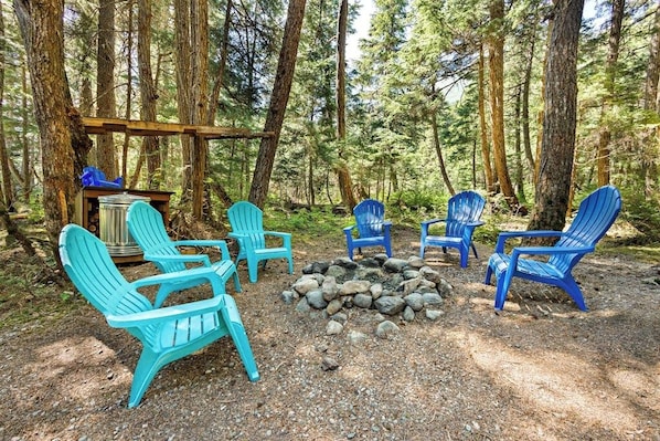 A forested campsite with several Adirondack chairs arranged around a fire pit.