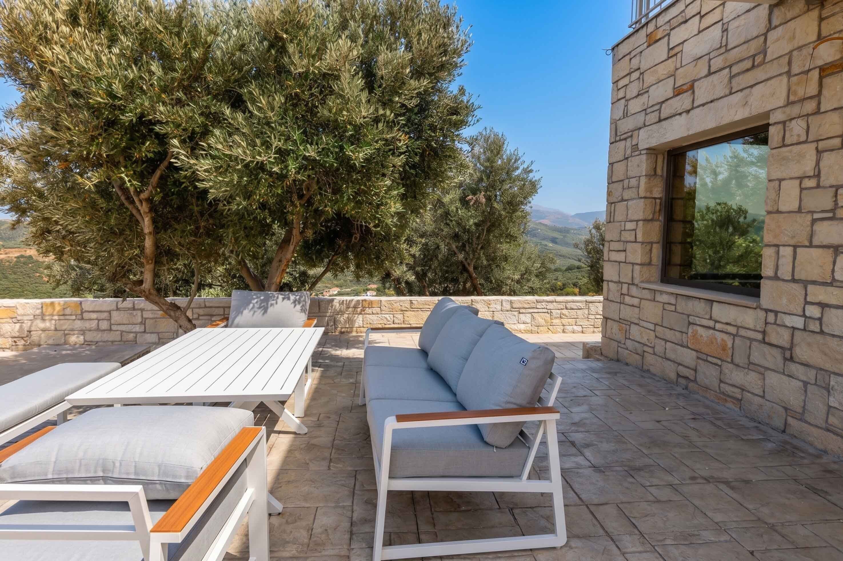 An outdoor seating area with a stone wall, a large tree, and a view of the countryside.